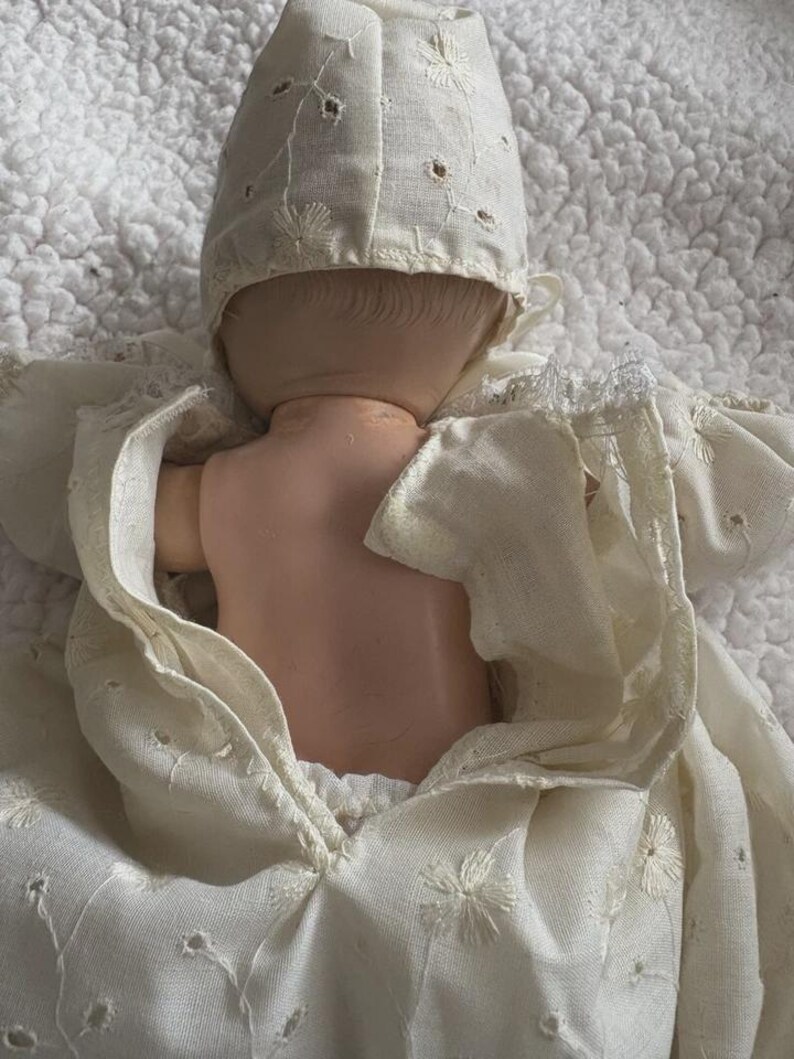 Op de afbeelding: Een vintage pop met een cr&egrave;mekleurige jurk en muts. De jurk en muts van de pop hebben delicate geborduurde bloempatronen en uitgesneden details. De huid van de pop is licht perzik van kleur.