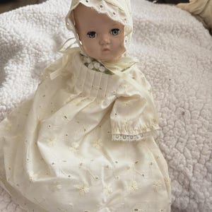 Op de afbeelding: Een vintage babypop met een cr&egrave;mekleurige oogjesjurk en muts. De pop heeft geschilderde gezichtskenmerken, waaronder blauwe ogen en een enigszins verdrietige uitdrukking. De jurk heeft bloemenborduurwerk en kanten afwerking.