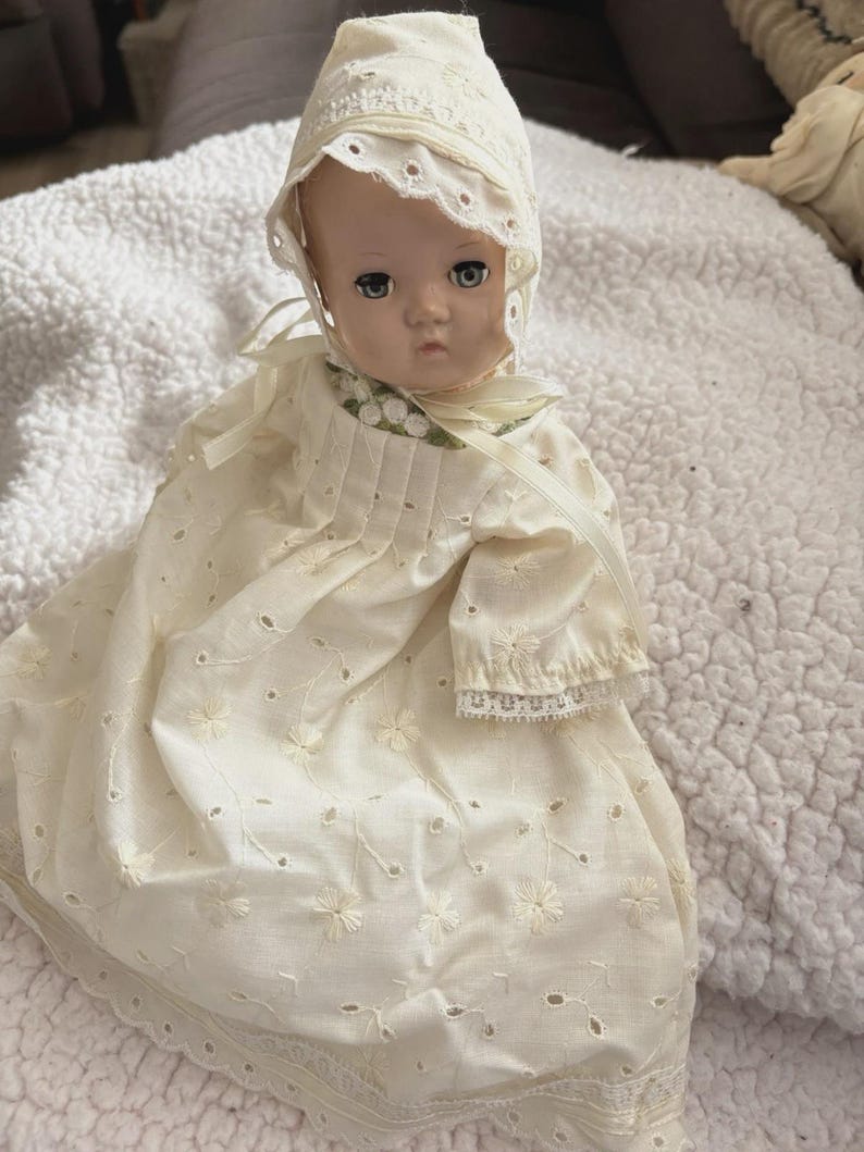 Op de afbeelding: Een vintage babypop met een cr&egrave;mekleurige oogjesjurk en muts. De pop heeft geschilderde gezichtskenmerken, waaronder blauwe ogen en een enigszins verdrietige uitdrukking. De jurk heeft bloemenborduurwerk en kanten afwerking.