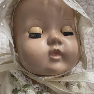 Op de afbeelding: Een vintage babypop met gesloten ogen en een cr&egrave;mekleurige muts. Het gezicht van de pop heeft een bleke teint met geschilderde kenmerken. De muts is afgezet met kant en een lint. De jurk van de pop is versierd met bloemenborduurwerk.