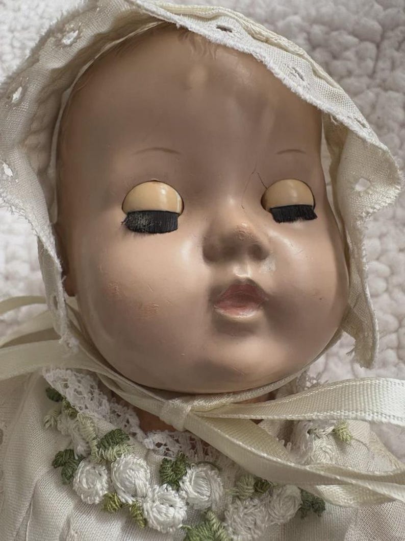 Op de afbeelding: Een vintage babypop met gesloten ogen en een cr&egrave;mekleurige muts. Het gezicht van de pop heeft een bleke teint met geschilderde kenmerken. De muts is afgezet met kant en een lint. De jurk van de pop is versierd met bloemenborduurwerk.