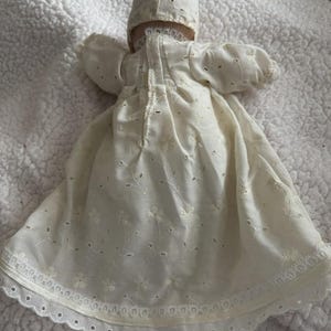 Op de afbeelding: Een vintage cr&egrave;mekleurige babypoppenjurk en muts. De jurk heeft delicate bloemenborduurwerk en oogjes, met een geschulpte kanten rand. De muts heeft een bijpassend ontwerp. De set is gemaakt van een zachte, lichte stof.