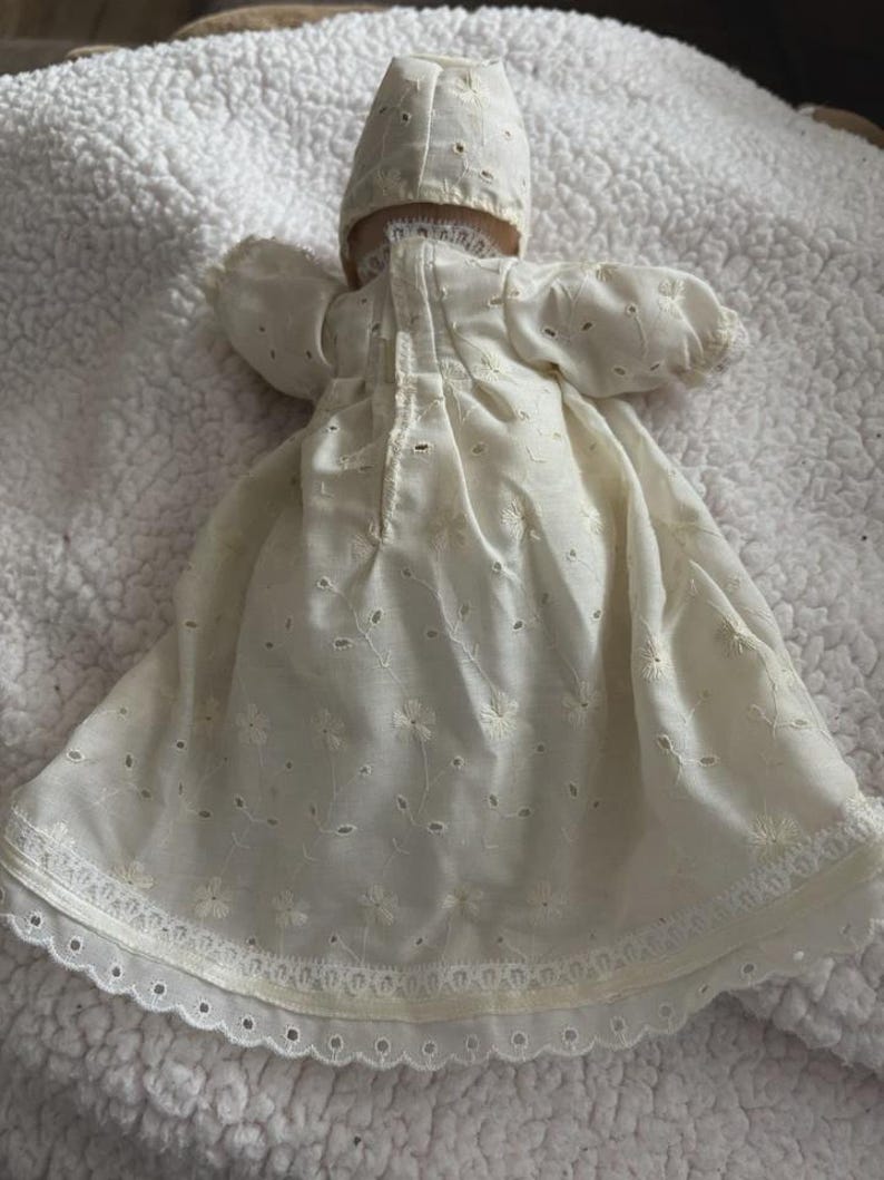 Op de afbeelding: Een vintage cr&egrave;mekleurige babypoppenjurk en muts. De jurk heeft delicate bloemenborduurwerk en oogjes, met een geschulpte kanten rand. De muts heeft een bijpassend ontwerp. De set is gemaakt van een zachte, lichte stof.