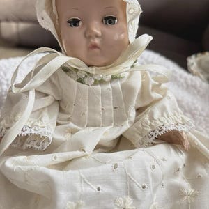 Op de afbeelding: Een vintage babypop met een witte geborduurde jurk en muts. De pop heeft een serene uitdrukking en draagt een cr&egrave;mekleurige outfit met bloemenborduurwerk en kanten randen. De muts is versierd met oogjes.