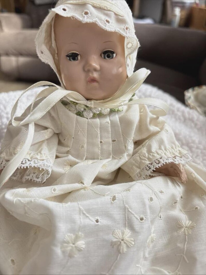 Op de afbeelding: Een vintage babypop met een witte geborduurde jurk en muts. De pop heeft een serene uitdrukking en draagt een cr&egrave;mekleurige outfit met bloemenborduurwerk en kanten randen. De muts is versierd met oogjes.