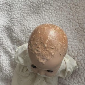 Op de afbeelding: Een vintage babypop met een geschilderd gezicht en gevormd haar. De pop draagt een witte jurk met oogjesdetails. Het hoofd van de pop is naar beneden gekanteld en de ogen zijn gesloten. De pop ligt op een wit, getextureerd oppervlak.
