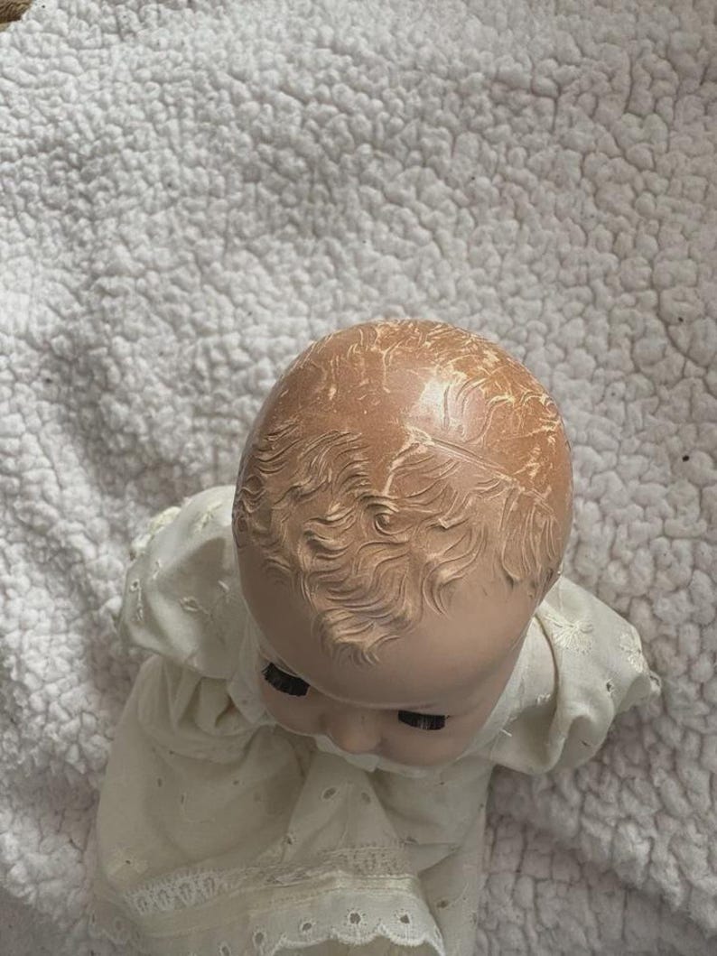 Op de afbeelding: Een vintage babypop met een geschilderd gezicht en gevormd haar. De pop draagt een witte jurk met oogjesdetails. Het hoofd van de pop is naar beneden gekanteld en de ogen zijn gesloten. De pop ligt op een wit, getextureerd oppervlak.