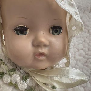 Op de afbeelding: Close-up van een vintage babypop met blauwe ogen en een cr&egrave;mekleurige muts. Het gezicht van de pop heeft zichtbare scheuren en ze draagt een witte jurk met bloemenborduurwerk. Een cr&egrave;mekleurig lint is om de nek geknoopt.
