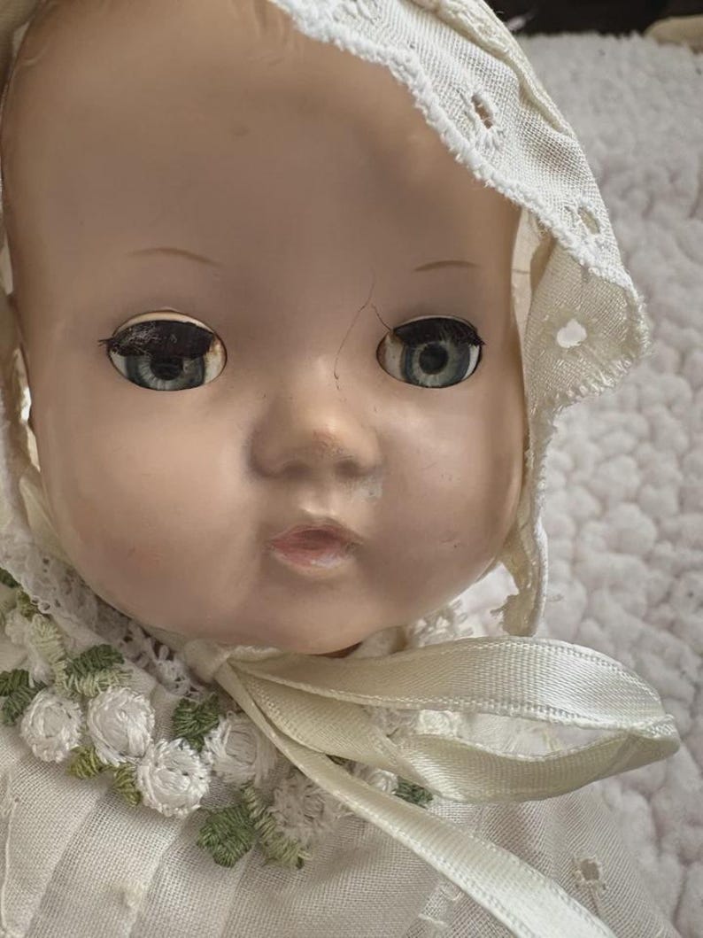 Op de afbeelding: Close-up van een vintage babypop met blauwe ogen en een cr&egrave;mekleurige muts. Het gezicht van de pop heeft zichtbare scheuren en ze draagt een witte jurk met bloemenborduurwerk. Een cr&egrave;mekleurig lint is om de nek geknoopt.