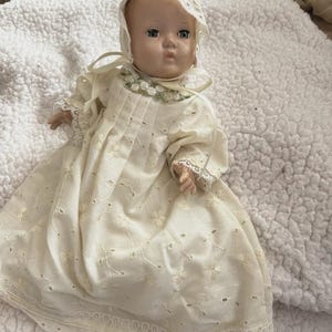 Op de afbeelding: Een vintage babypop met een cr&egrave;mekleurige jurk en muts. De jurk heeft bloemenborduurwerk en kanten randen. De pop heeft blauwe ogen en een serene uitdrukking, die een gevoel van nostalgie oproept.