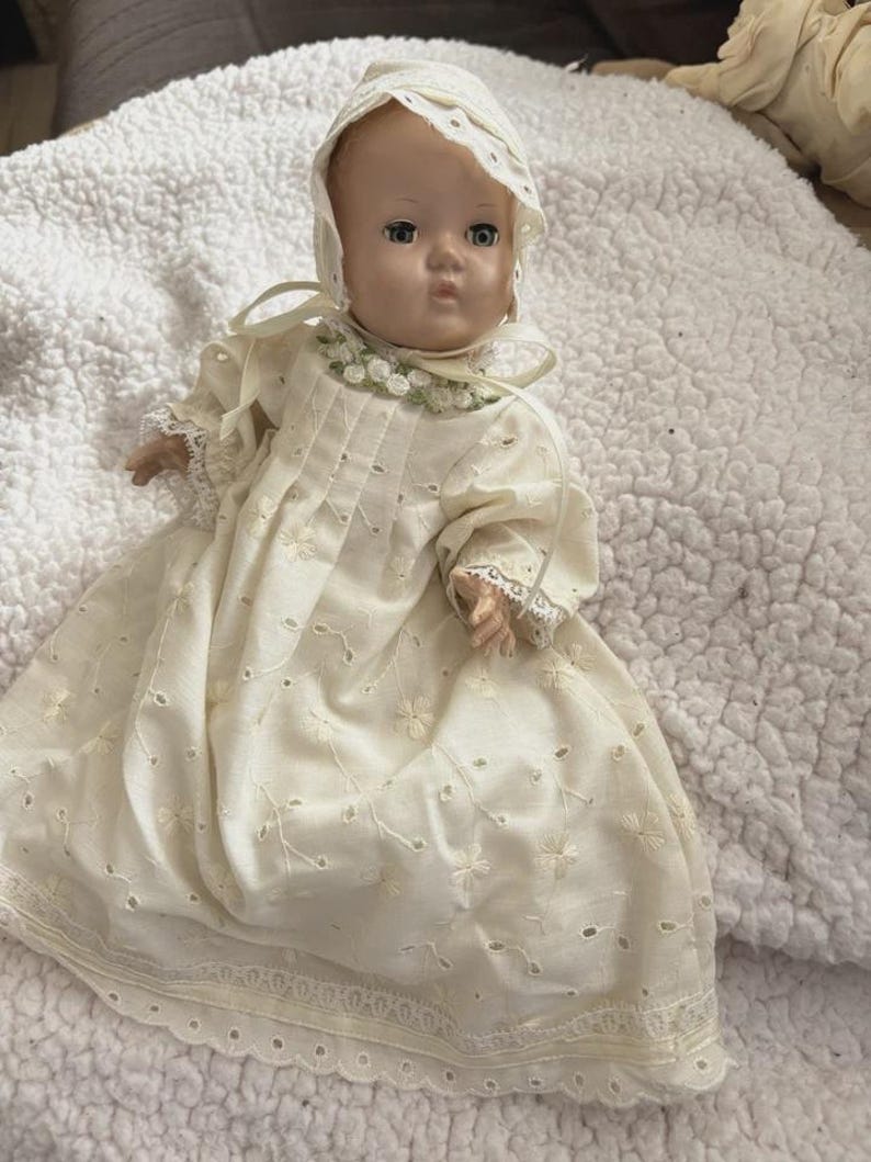Op de afbeelding: Een vintage babypop met een cr&egrave;mekleurige jurk en muts. De jurk heeft bloemenborduurwerk en kanten randen. De pop heeft blauwe ogen en een serene uitdrukking, die een gevoel van nostalgie oproept.