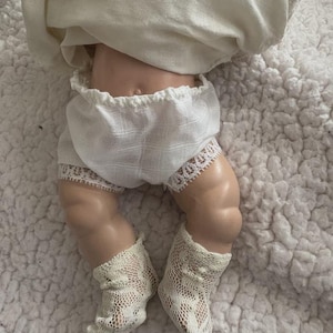 Op de afbeelding: Een vintage pop met een porseleinen lichaam, gekleed in een witte, met kant afgezette bloomer, een witte top en delicate kanten sokken. De benen van de pop zijn zichtbaar en tonen het gedetailleerde vakmanschap van het antieke speelgoed. De pop ligt op een wit, getextureerd oppervlak.