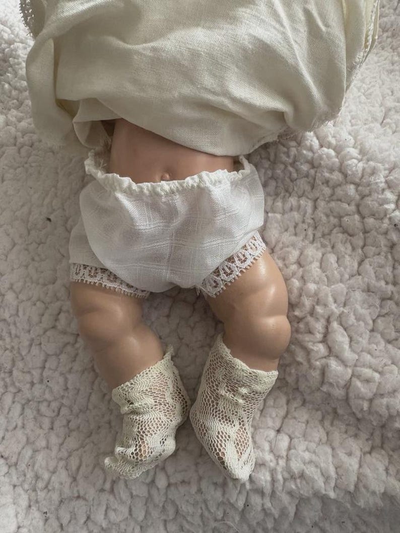 Op de afbeelding: Een vintage pop met een porseleinen lichaam, gekleed in een witte, met kant afgezette bloomer, een witte top en delicate kanten sokken. De benen van de pop zijn zichtbaar en tonen het gedetailleerde vakmanschap van het antieke speelgoed. De pop ligt op een wit, getextureerd oppervlak.