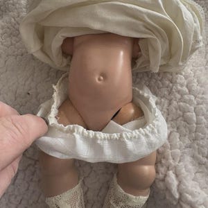 Op de afbeelding: Een vintage pop met een porseleinen torso en benen, gekleed in een witte jurk en kanten sokken. De huid van de pop is licht perzikkleurig en de jurk heeft een gerimpelde rok. De benen van de pop zijn gebogen bij de knie&euml;n.