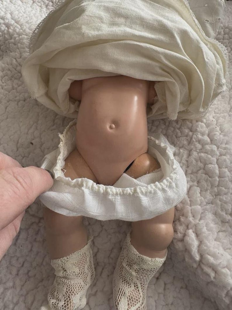 Op de afbeelding: Een vintage pop met een porseleinen torso en benen, gekleed in een witte jurk en kanten sokken. De huid van de pop is licht perzikkleurig en de jurk heeft een gerimpelde rok. De benen van de pop zijn gebogen bij de knie&euml;n.