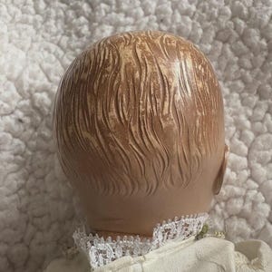 Op de afbeelding: Een vintage poppenhoofd, gezien van achteren, met gevormd, getextureerd haar in lichtbruine en bruine tinten. De pop draagt een witte jurk met een kanten kraag. De achtergrond is een getextureerd, wit oppervlak.