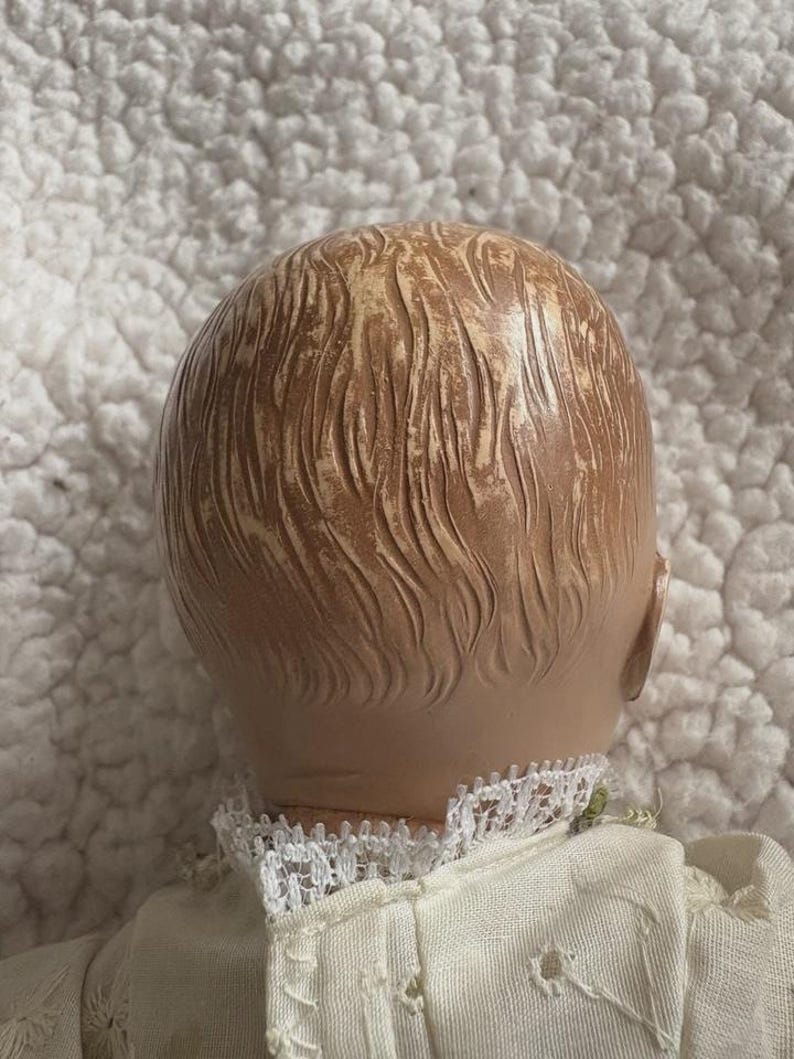 Op de afbeelding: Een vintage poppenhoofd, gezien van achteren, met gevormd, getextureerd haar in lichtbruine en bruine tinten. De pop draagt een witte jurk met een kanten kraag. De achtergrond is een getextureerd, wit oppervlak.