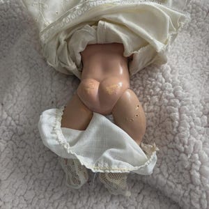 Op de afbeelding: Een vintage pop met een porseleinen lichaam en een witte jurk. De benen van de pop zijn zichtbaar, met witte kanten sokken. De jurk heeft geborduurde details en een gefranjerde zoom. De pop ligt op een wit, getextureerd oppervlak.