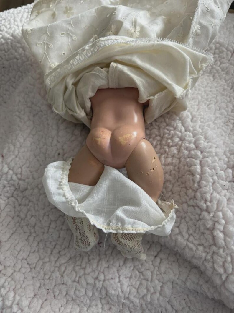Op de afbeelding: Een vintage pop met een porseleinen lichaam en een witte jurk. De benen van de pop zijn zichtbaar, met witte kanten sokken. De jurk heeft geborduurde details en een gefranjerde zoom. De pop ligt op een wit, getextureerd oppervlak.