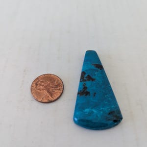 Peut inclure: Une pierre précieuse triangulaire turquoise polie avec des inclusions brun foncé. Une pièce de monnaie est à côté de la pierre précieuse pour la comparaison de taille. La pierre précieuse a une finition lisse et brillante.