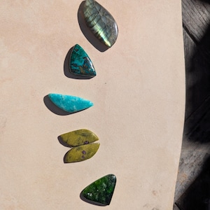 May include: An assortment of polished gemstones in various shapes and colors. The collection includes a teardrop-shaped iridescent stone, a triangular teal stone, a turquoise stone, two olive-green stones, and a dark green triangular stone.