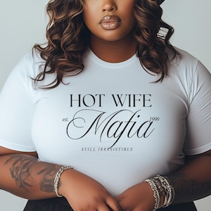 May include: White t-shirt with the text "HOT WIFE Mafia" in black script. The shirt includes the text "est. 1999" and "STILL IRRESISTIBLE" in a simple font. The shirt is a crew neck style.