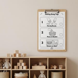 May include: A wooden clipboard featuring a white instructional chart titled Putting Away Toys. The chart displays three numbered steps with black line illustrations: 1. Pick Up Blocks, 2. Place in Basket, and 3. Return to Shelf, each with a checkbox.