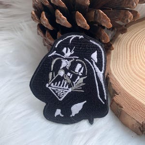 Könnte beinhalten: Schwarz-weißer, gestickter Aufnäher mit Darth Vaders Helm und Gesicht. Der Aufnäher ist eine detaillierte Darstellung der ikonischen Figur aus dem Star Wars-Franchise. Der Aufnäher ist zum Aufbügeln oder Aufnähen auf Kleidung oder Accessoires gedacht.