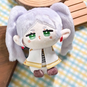 May include: A plush doll with light lavender hair styled in two high pigtails, embroidered green eyes, and pointed ears. The doll wears a white jacket with gold trim and red accents, featuring a black and white striped shirt underneath and brown shoes.