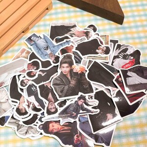 May include: A collection of die-cut stickers featuring various photographic portraits of a person with dark hair in different outfits, including a dark robe, a denim jacket, and a patterned tunic, with some stickers showing the person holding a small object.