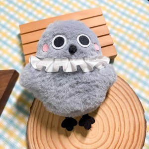 Op de afbeelding: Een knuffel in de vorm van een ronde, grijze vogel met een zachte, pluizige textuur. Het heeft twee grote witte ogen met zwarte pupillen, een kleine zwarte snavel en twee roze cirkelvormige vlekken op de wangen. Een witte ruchekraag zit om de nek.