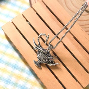 May include: A silver-toned metal pendant necklace featuring a detailed, ornate oil lamp design with a curved handle and spout. The surface of the lamp is decorated with intricate, embossed scrollwork patterns and a small, red accent stone.