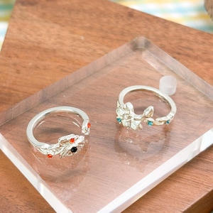 May include: Two silver-toned open rings featuring floral motifs. The left ring has orange and black stones, while the right ring features a clear center stone with small blue accents. Both rings have a polished metallic finish and adjustable bands.
