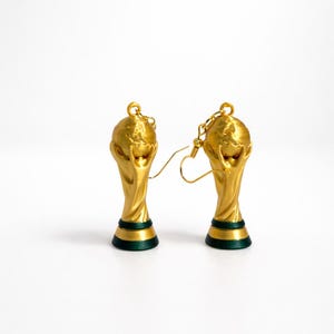 May include: A pair of gold-colored earrings shaped like the FIFA World Cup trophy. The earrings have a gold finish with a green and black base. The earrings are on a white background.