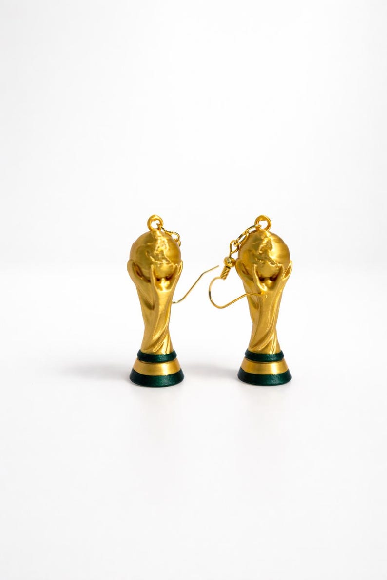 May include: A pair of gold-colored earrings shaped like the FIFA World Cup trophy. The earrings have a gold finish with a green and black base. The earrings are on a white background.