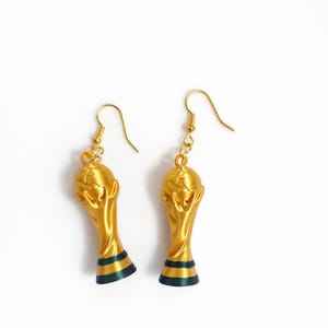 May include: A pair of gold-colored trophy earrings with a dark blue and green stripe at the base. The earrings are suspended from gold-colored ear wires, set against a white background.