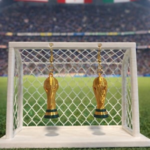 May include: Gold-colored trophy earrings with a black base, hanging in front of a miniature soccer goal. The background features a soccer field and flags of the USA, Canada, and Mexico. The earrings are a sports-themed accessory.