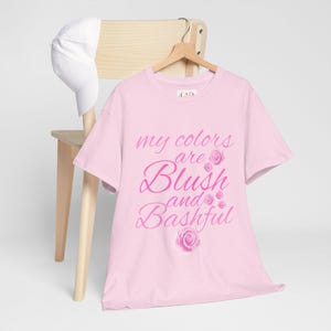 May include: A pale pink t-shirt featuring the text "my colours are Blush and Bashful" in a cursive font, with rose illustrations. A white baseball cap is on a wooden chair behind the shirt.