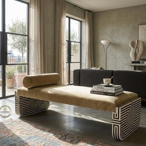 Peut inclure: Une chaise longue beige avec des pieds à motifs géométriques. La chaise longue a un coussin cylindrique et se trouve dans un salon moderne. Des livres et une tasse à thé sont posés sur la chaise longue. La pièce a de grandes fenêtres et une décoration aux tons neutres.
