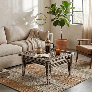 May include: A square coffee table featuring a dark brown and white geometric inlay pattern on its surface and four tapered legs. The table holds a glass French press, a ceramic mug, an open book, and a small orange candle in a glass jar.