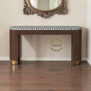 May include: A brown console table with a chequered top and ribbed sides. The table has a gold-coloured base and is positioned beneath an ornate, oval-shaped mirror. The table's design features a mix of textures and patterns.