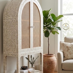 Può includere: Un mobile bianco con la parte superiore ad arco e ante in canna intrecciata. Il mobile presenta un motivo a foglie e un ripiano inferiore. La stanza comprende una pianta in un vaso di terracotta e una poltrona beige.
