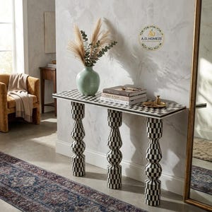 May include: A rectangular console table featuring a black and white chequered pattern on the top surface and three stacked, cylindrical, chequered legs. A light green vase with dried foliage and a stack of books sit on the table surface.