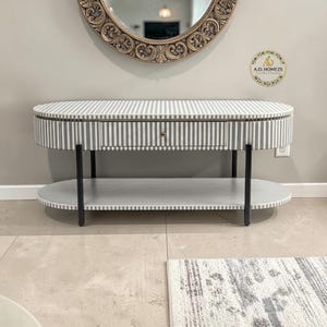 May include: Oval-shaped console table with a gray and white striped pattern. The table has a drawer with a gold-colored knob and a lower shelf. Black legs support the table. A decorative mirror hangs above the table.