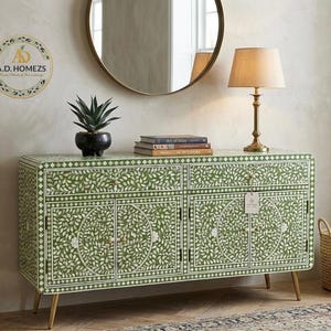 May include: Green and white floral patterned cabinet with gold legs. The cabinet has two doors and two drawers. A round gold mirror hangs above the cabinet. A lamp, a plant, and books are on top of the cabinet. A woven basket sits on the floor.
