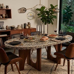 Peut inclure: Une table de salle à manger ovale avec un plateau à motifs et une base en bois est dressée pour un repas. La table est entourée de chaises en bois avec des sièges en cuir noir. Un lustre moderne est suspendu au-dessus de la table.