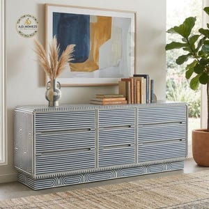May include: A rectangular sideboard featuring a blue and white striped pattern with rounded corners and six drawers. The base has a curved geometric design. A ceramic vase with dried pampas grass and a row of books sit on the top surface.