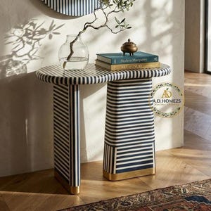 Könnte beinhalten: Ein Beistelltisch mit einem schwarz-weiß gestreiften Design. Der Tisch hat eine runde Oberseite und eine rechteckige Basis mit goldenen Akzenten. Eine Glasvase mit einem Zweig, Büchern und einem kleinen Behälter befindet sich auf dem Tisch.
