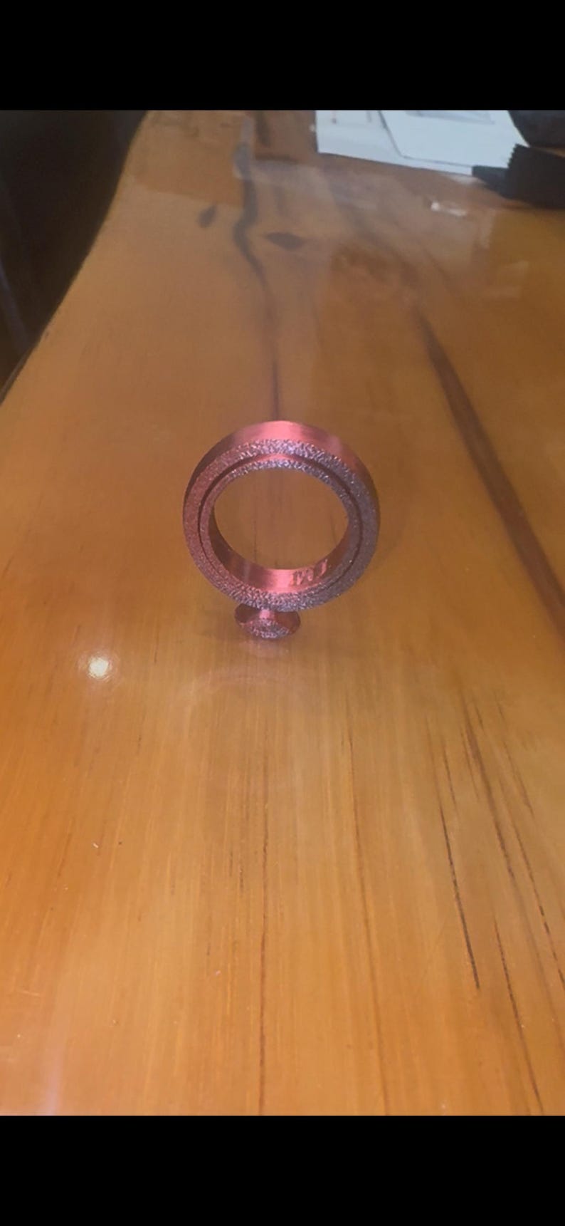 Pu&ograve; includere: Un oggetto a forma di anello marrone rossiccio con una superficie strutturata, appoggiato su una superficie di legno. L'anello ha una piccola base e un'apertura centrale. L'oggetto sembra essere un oggetto decorativo o un gioiello.