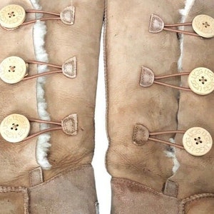 May include: A pair of brown suede boots with four gold buttons on each boot. The boots have a sheepskin lining and a rubber sole.